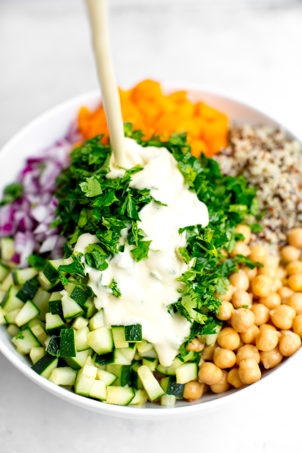 Creamy feta dressing being poured onto ingredients for mediterranean quinoa salad made with orange bell pepper, red onions, parsley, tricolored quinoa, cucumbers and chickpeas