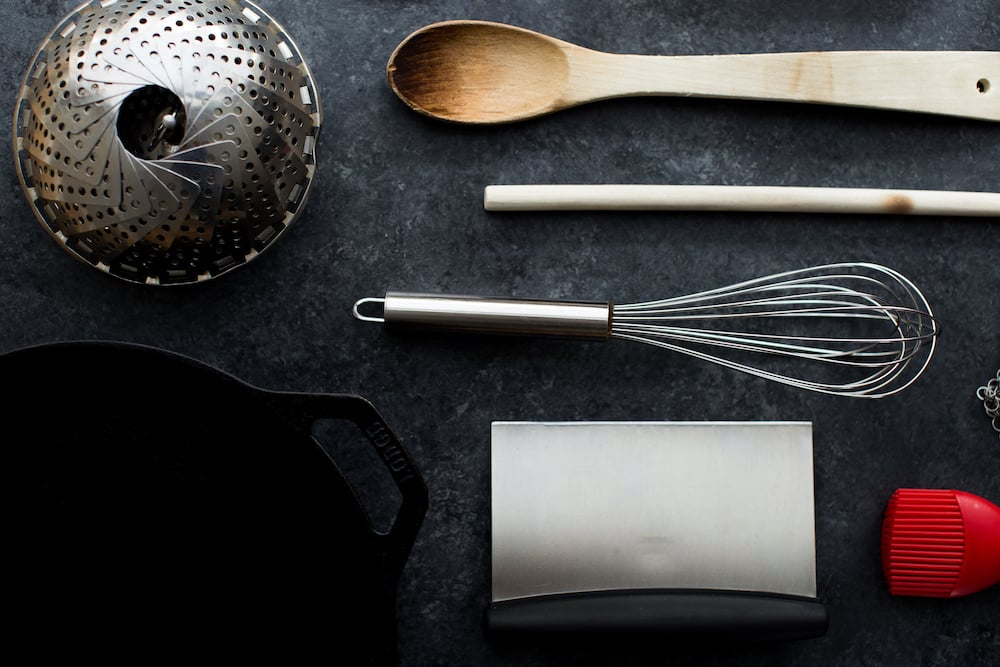 kitchen equipment including whisk steamer wooden spoon bench scraper and cast iron skillet on dark background