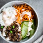 overhead view of healthy korean bulgogi with beef, rice, carrots, cucumber and sriracha crema in gray ceramic bowl with gray background napkin and fork