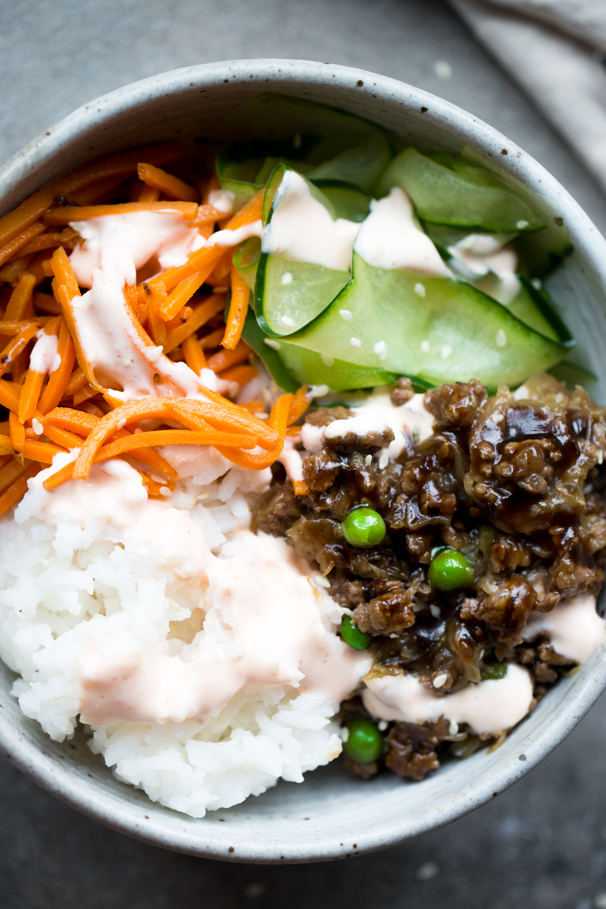 overhead view of healthy korean bulgogi with beef, cabbage, peas, rice, carrots, cucumber and sriracha crema in gray ceramic bowl with gray background