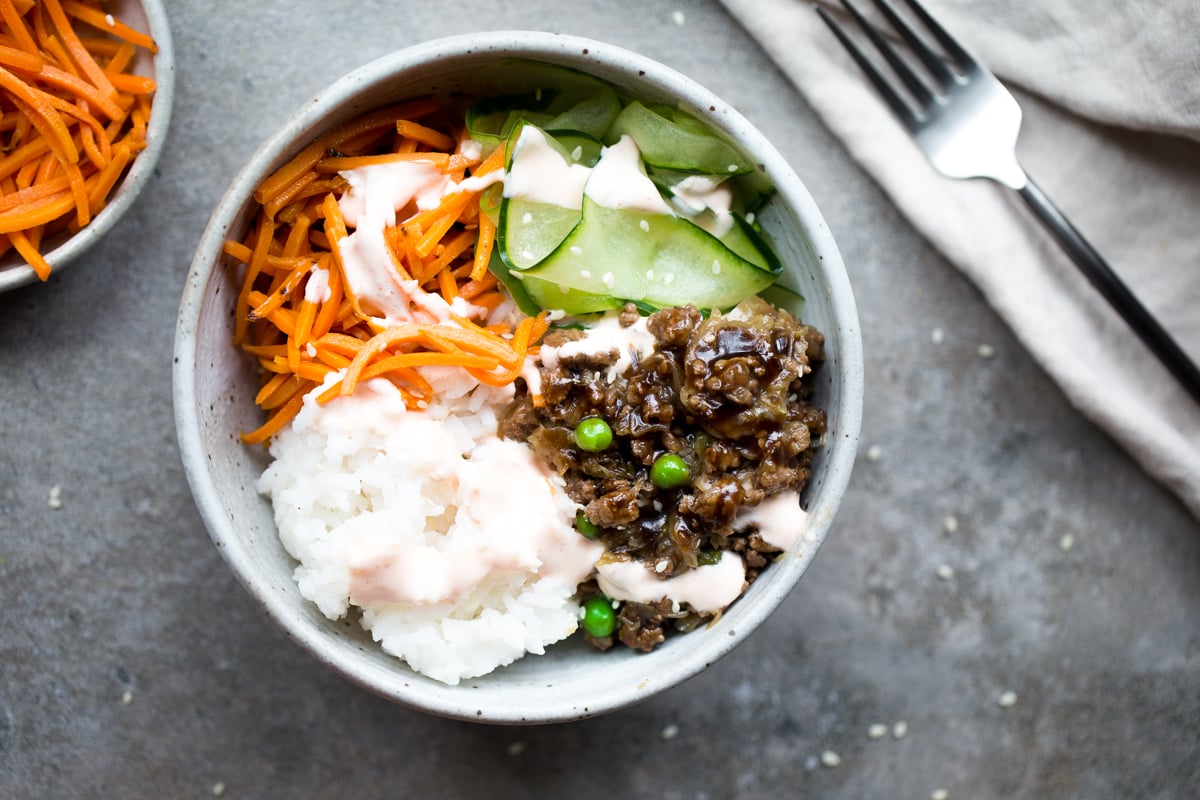 overhead horizontal photo of ground beef, cabbage, peas, rice, carrots, cucumber and sriracha crema in gray ceramic bowl with gray background