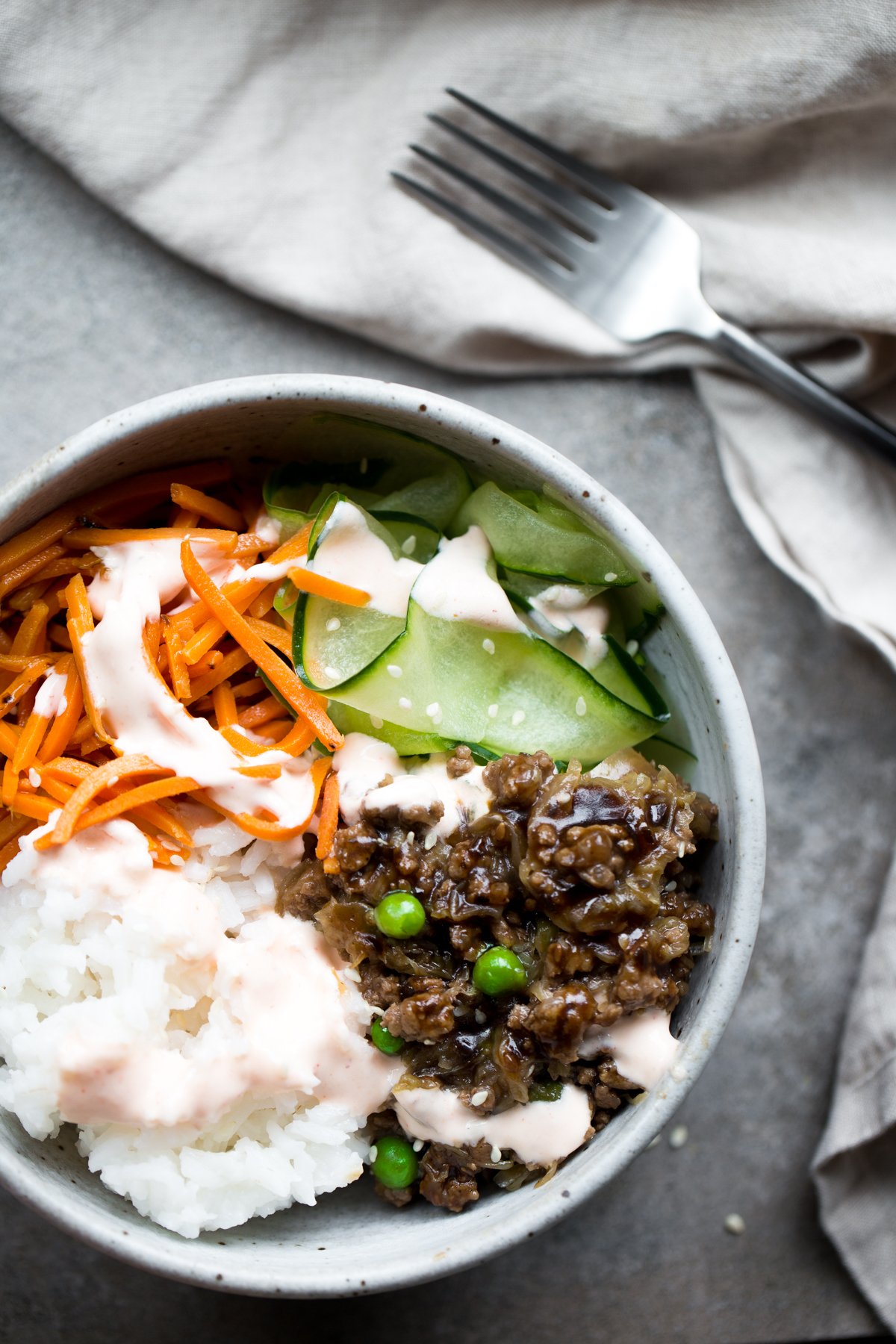 overhead view of korean bulgogi with beef, rice, carrots, cucumber and sriracha crema in gray ceramic bowl with gray background napkin and fork