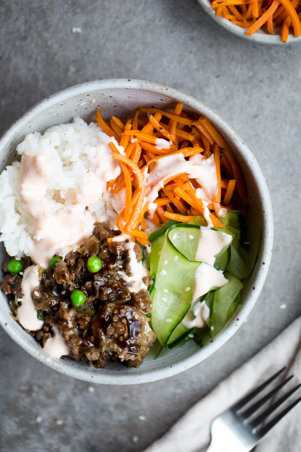 overhead view of healthy korean bulgogi with beef, rice, carrots, cucumber and sriracha crema in gray ceramic bowl with gray background napkin and fork