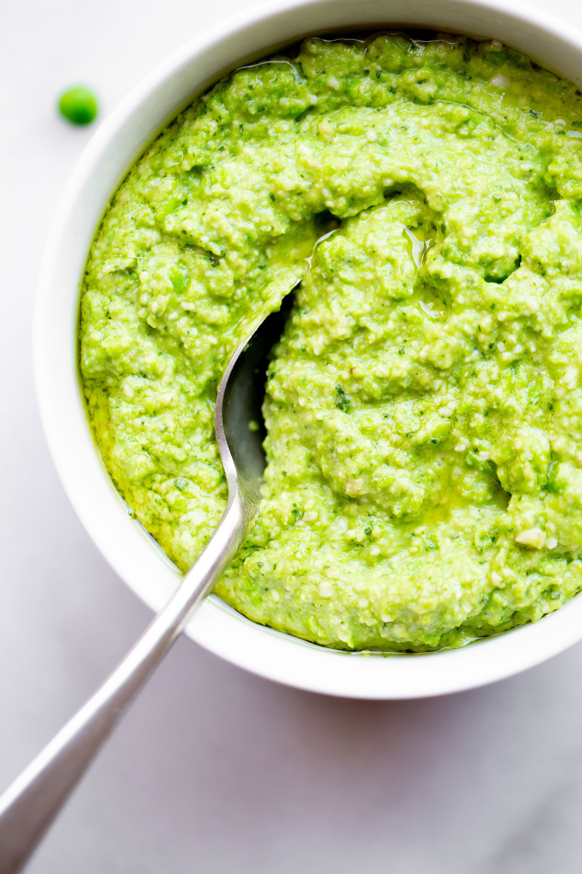 overhead of a bowl of pea pesto with a spoon on a white marble background