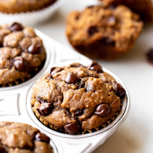 closeup photo at 45 degree angle of freshly baked healthy chocolate chip banana muffins in an aluminum muffin pan on a light gray marble background