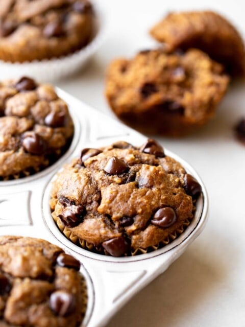 closeup photo at 45 degree angle of freshly baked healthy chocolate chip banana muffins in an aluminum muffin pan on a light gray marble background