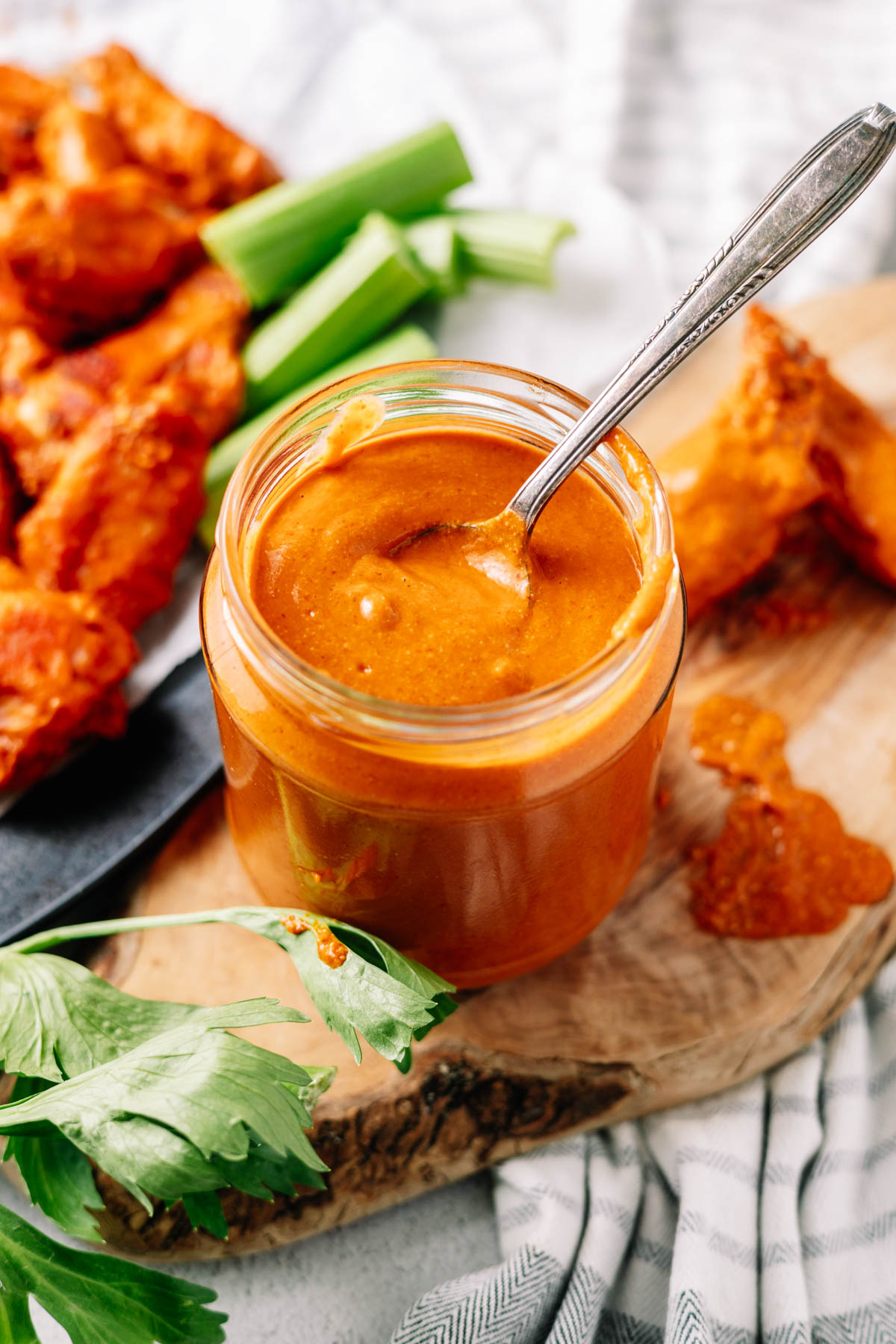 A glass jar of thick Buffalo sauce with a spoon and wings with celery in the background