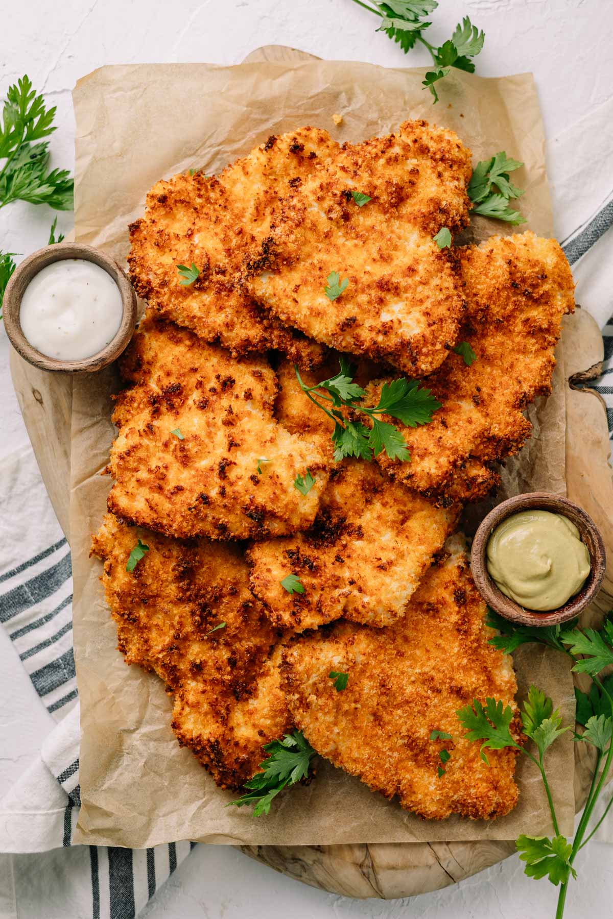 A plate of crispy, golden air fryer chicken cutlets on brown paper with dipping sauce, garnished with parsley, on white background