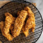 Overhead photo of golden, crispy air fryer chicken tenders on a wire rack. The marble background contains the air fryer basket in the corner and a small bowl of honey mustard sauce