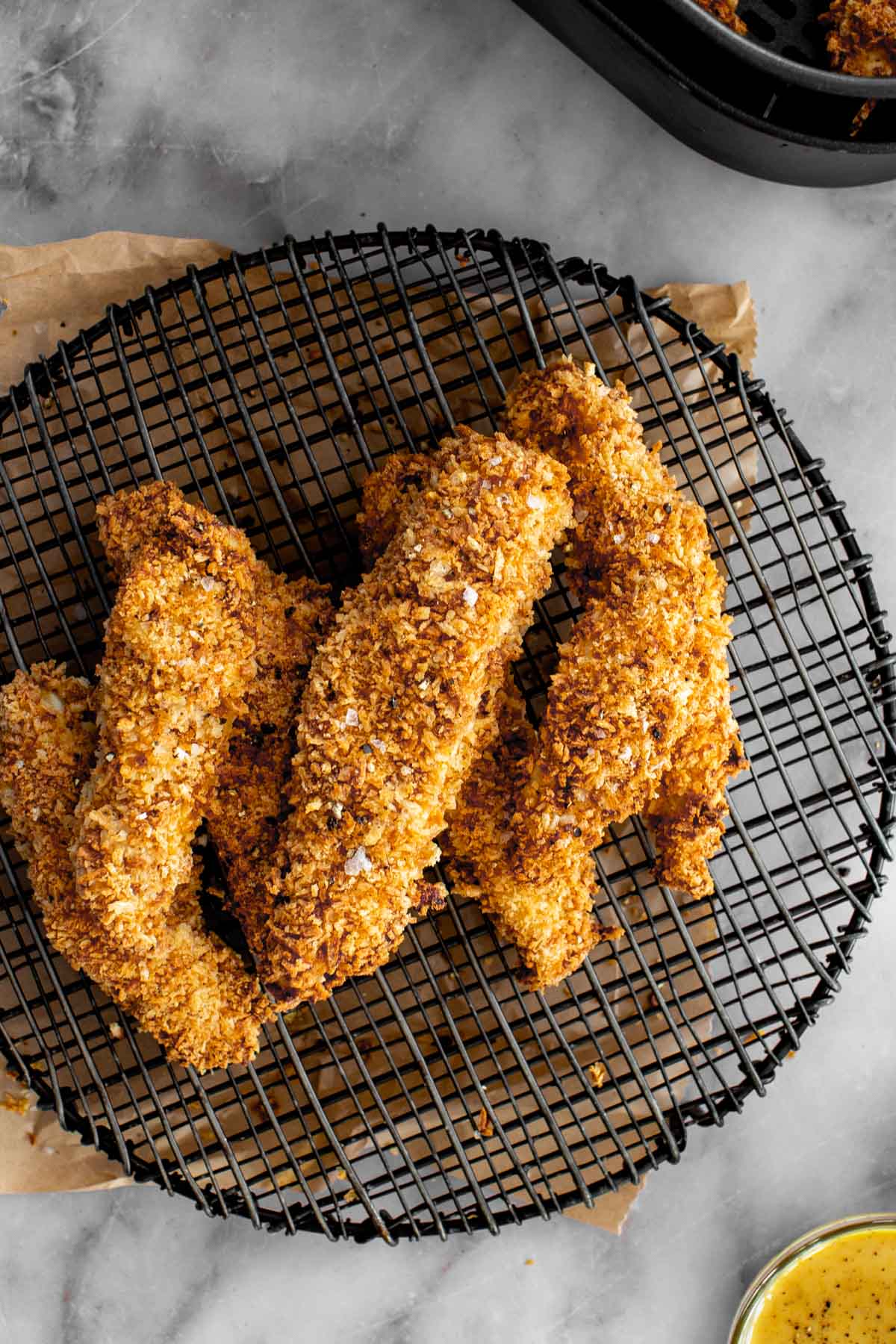 Overhead photo of golden, crispy air fryer chicken tenders on a wire rack. The marble background contains the air fryer basket in the corner and a small bowl of honey mustard sauce