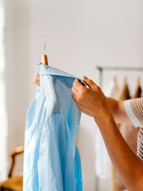 Close-up of a person examining a light blue button-down shirt on a wooden hanger, symbolizing intentional wardrobe choices and mindful shopping habits.