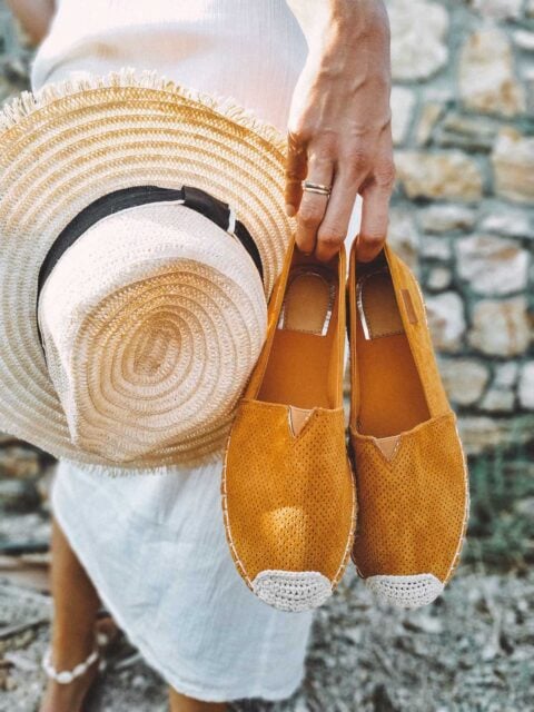 Woman in a white linen dress from a summer capsule wardrobe holding mustard yellow espadrilles and a straw sun hat