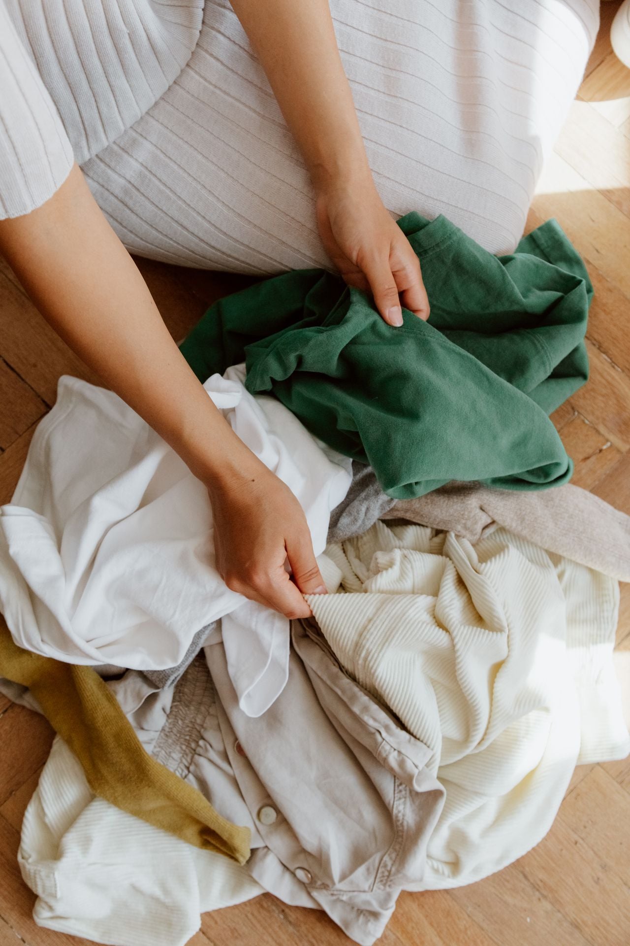 Hands sorting through a pile of clothes on a wood floor while decluttering a closet